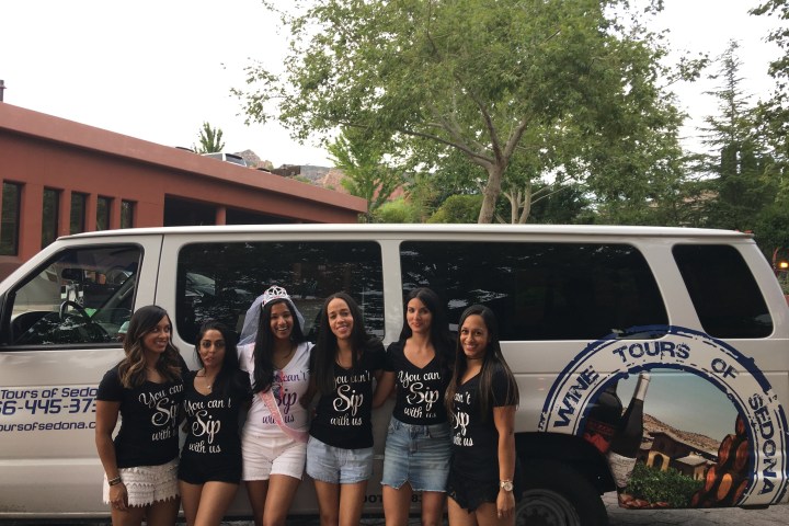 Six women in matching shirts stand in front of a tour van, trees and building in the background.