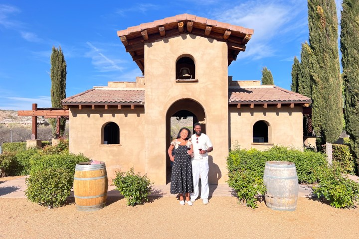 Two people standing in front of a rustic house with wine barrels and trees.