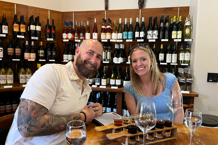 Two people smiling at a wine tasting with bottles on shelves in the background.