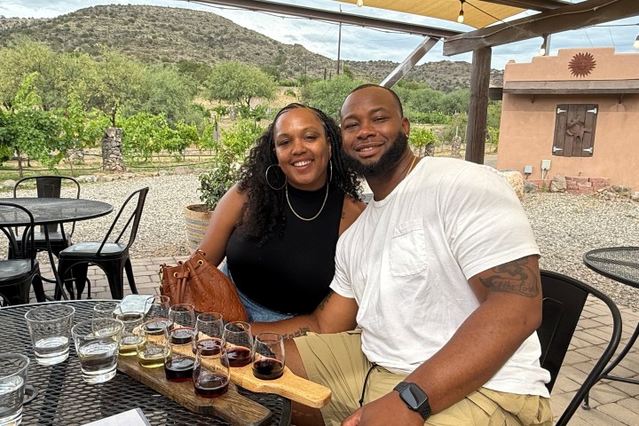 Smiling couple sitting at outdoor winery table with wine flights, vineyard and hills in background.