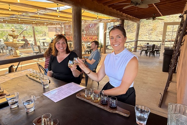 Two women sitting at a table outdoors, smiling and holding drinks, with tasting trays and water glasses.