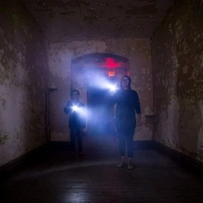 Two people exploring dark hallway with flashlights, exit sign above.