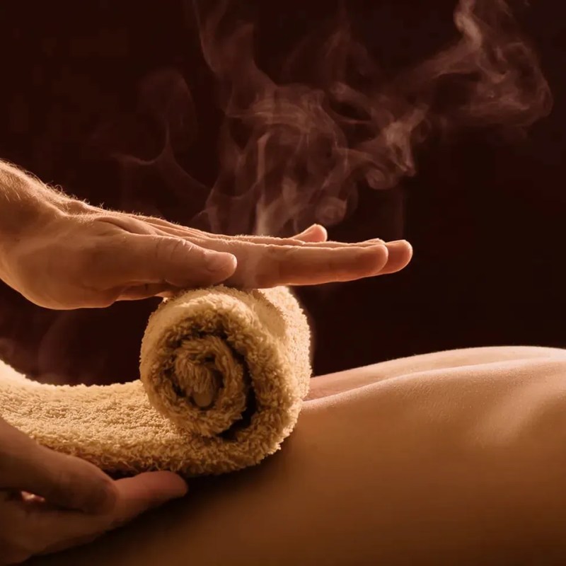 Hands holding steaming rolled towel over a person's back against a dark background.