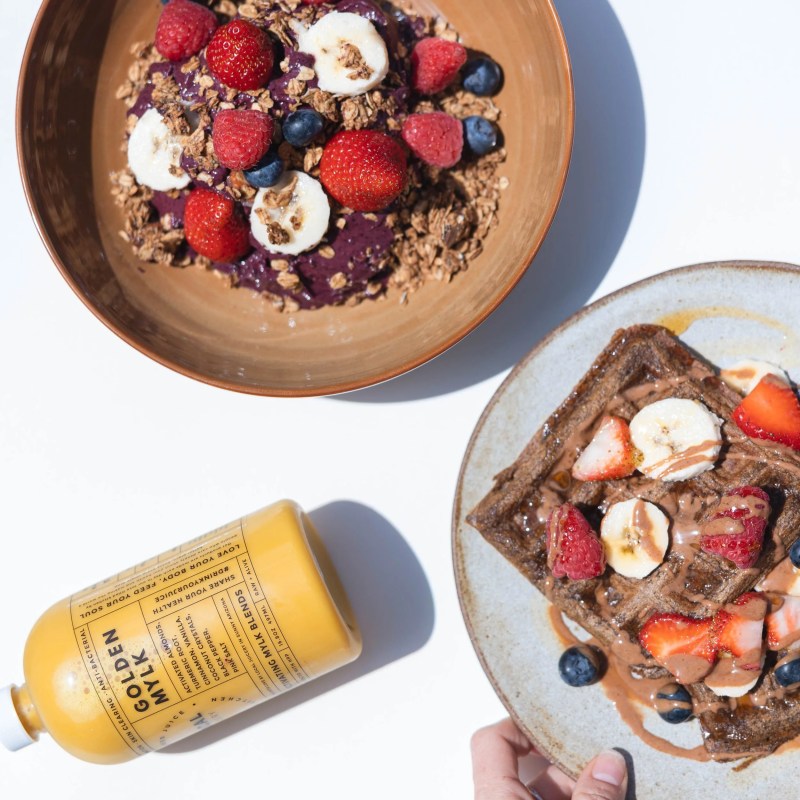 Acai bowl with berries, waffles with fruit, and a bottle of golden milk on a white background.