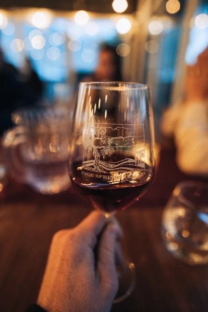 Hand holding wine glass with Page Springs logo, blurred background.