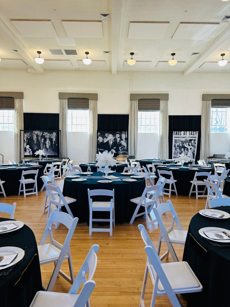 Decorated dining hall with round tables, black tablecloths, white chairs, and vintage photos on display.