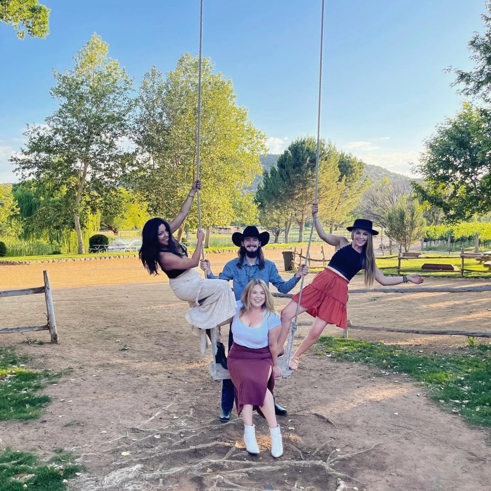 Four people on swings in a sunny park, two standing, two sitting, with trees and hills in the background.