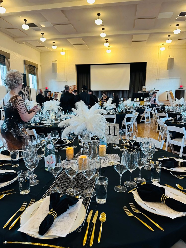 Formal event with round tables, gold utensils, glasses, and white feathers as centerpieces.