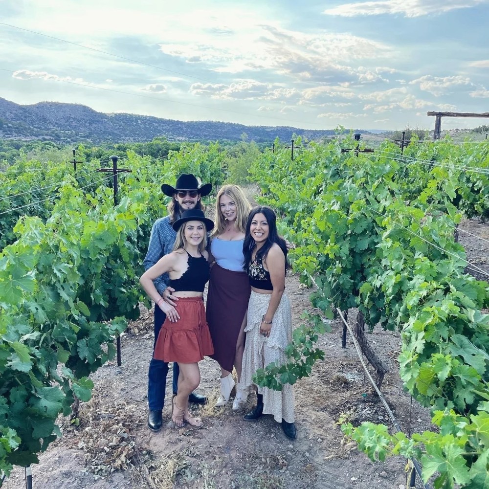 Four people standing in a vineyard with green grapevines and hills in the background.