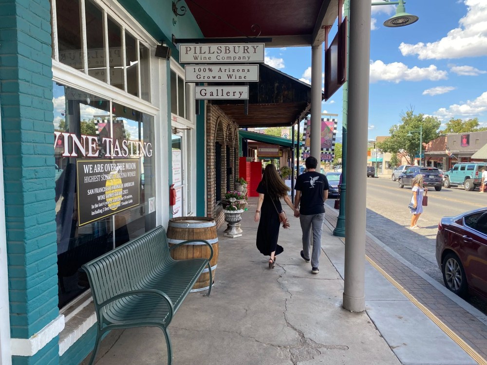 A couple walks past a wine tasting shop on a sunny street with benches and cars.