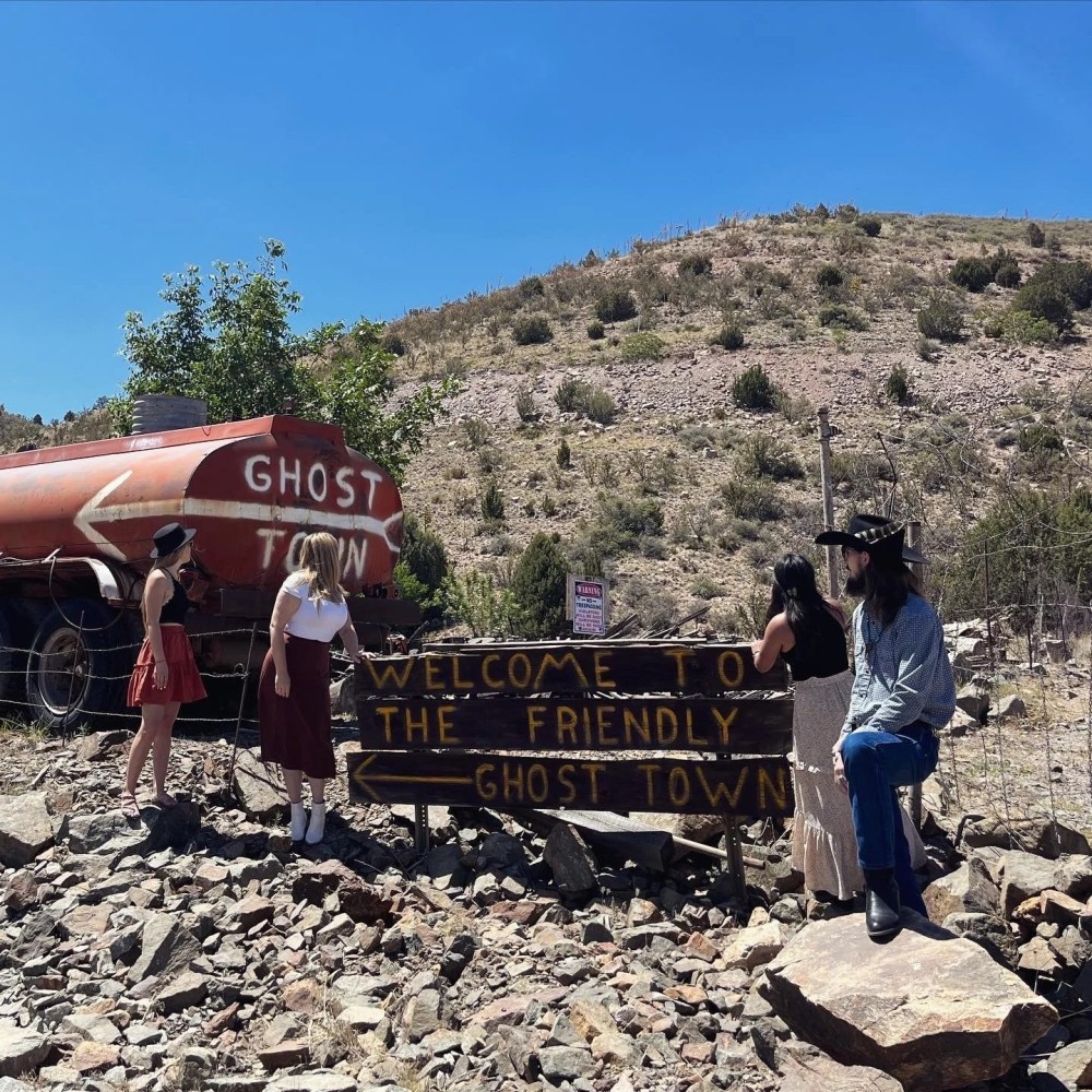 People at ghost town sign near old tanker in rocky desert landscape.