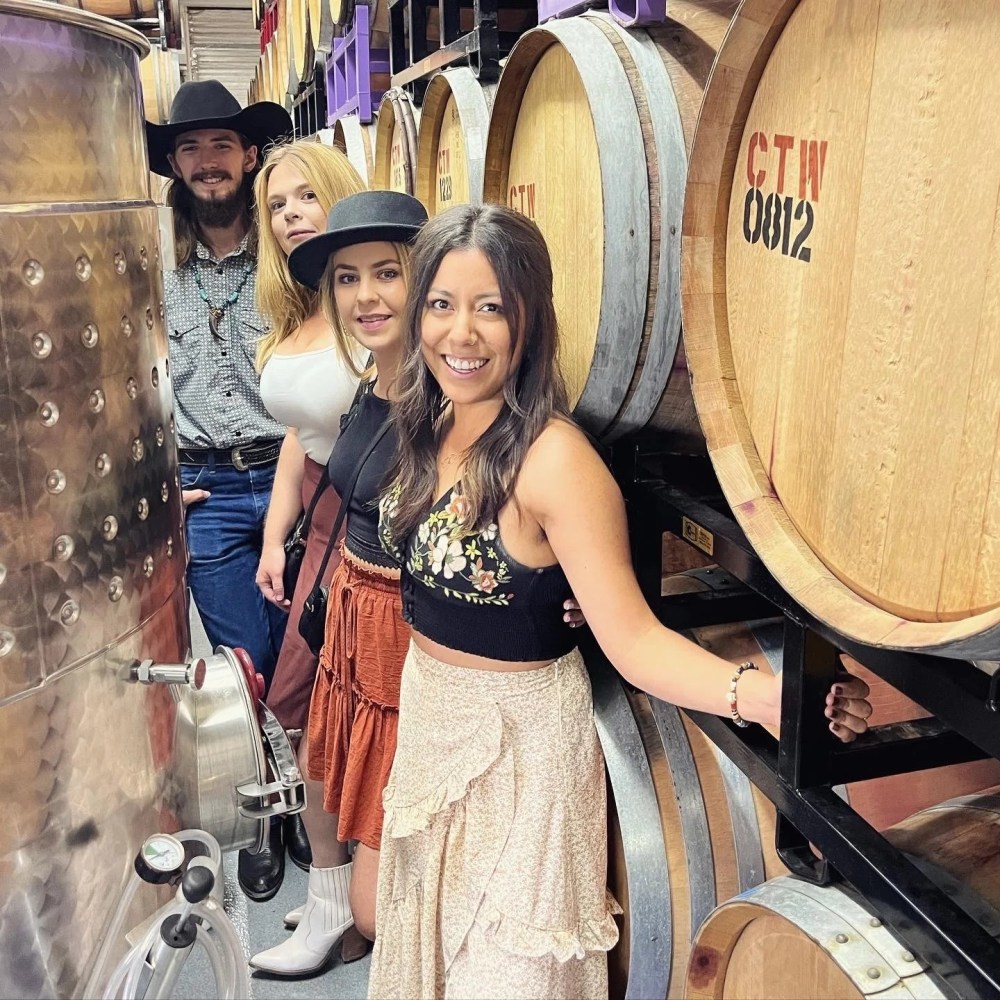 Four people smiling in a winery, standing beside large wooden barrels and a metal tank.