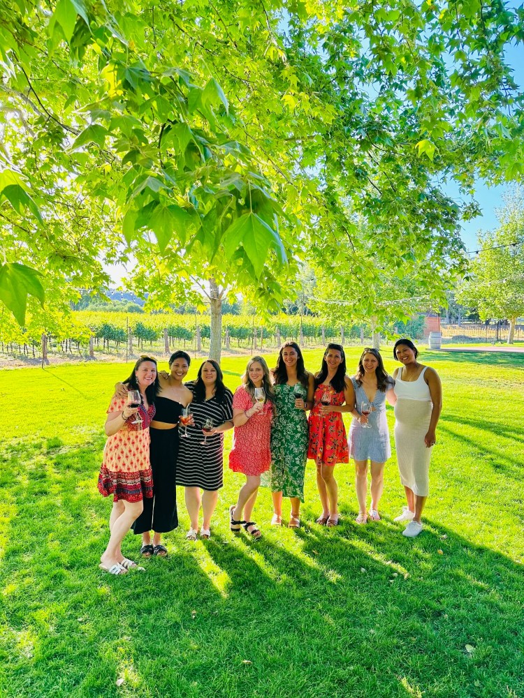 Seven women in dresses stand under a tree holding wine glasses in a sunny vineyard.