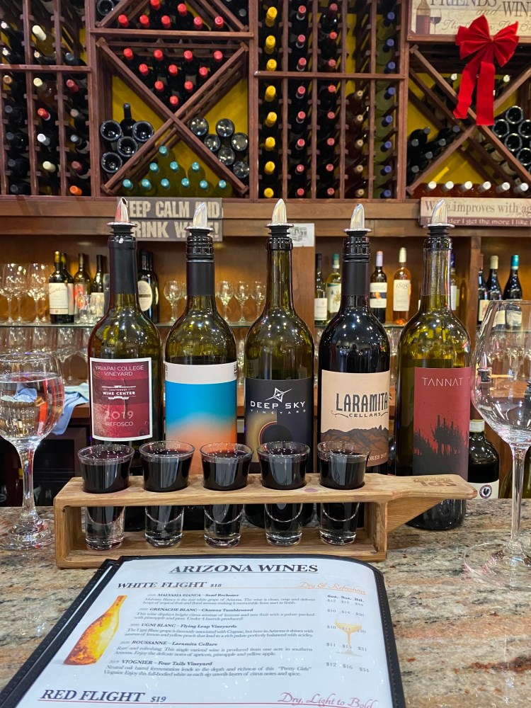 Five wine bottles with a flight of glasses on a bar with wine racks in the background.