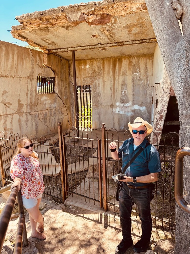 Two people near a rusty metal gate, by a crumbling concrete structure under a clear sky.