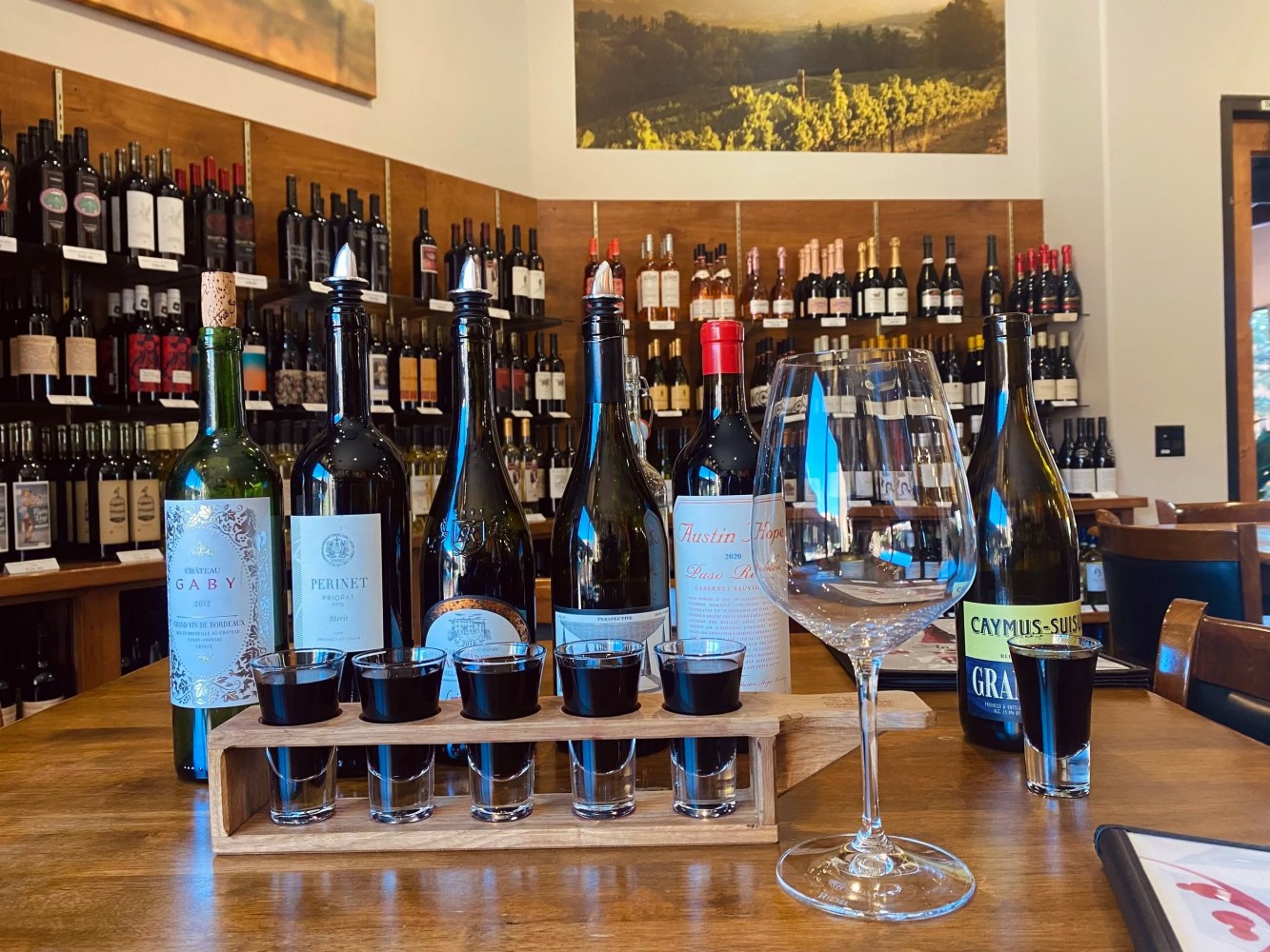 Wine bottles and flight on a table in a tasting room with wooden shelves filled with bottles.